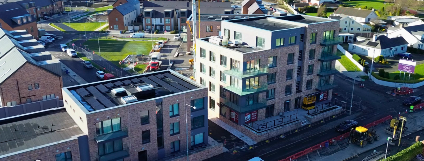 Aerial view of a newly completed mid rise apartment development with brick facades, balconies and ground floor commercial units, set within an active residential neighbourhood with surrounding housing, roads and ongoing construction works visible.